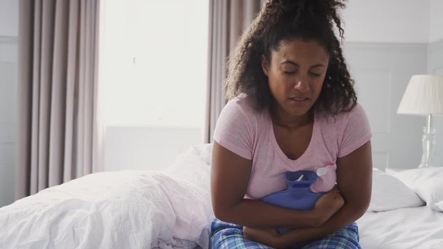 Woman Wearing Pajamas Suffering With Period Pain Sitting On Bed With Hot Water Bottle