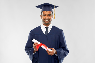 education, graduation and people concept - happy smiling indian male graduate student in mortar board and bachelor gown with diploma over grey background