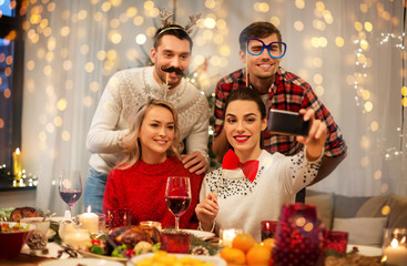 Obraz na płótnie Canvas holidays and celebration concept - happy friends with party props taking selfie by smartphone at home christmas dinner