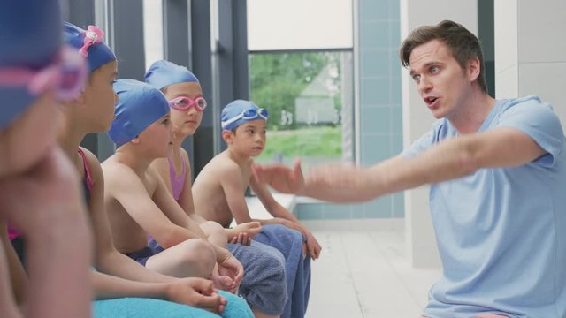 Male Coach Giving Children In Swimming Class Briefing As They Sit On Edge Of Indoor Pool