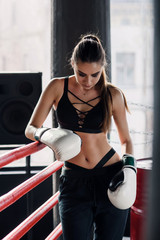 Fototapeta na wymiar Tired sports girl leaned on red ropes on boxing ring, and have a rest after hard training in black loft gym. Healthy and sporty lifestyle concept.