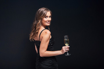 Young woman in elegant dress staying with glass of champagne against black background.