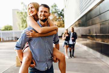 Young excited friends are having fun in the city, students are walking, in the foreground a man carries a girl on his back