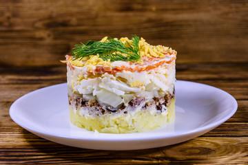Plate with traditional russian salad Mimosa on a wooden table. Layered salad with potatoes, sardine, cheese, carrot, eggs and mayonnaise