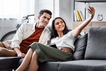 happy multicultural students smiling and taking selfie in apartment