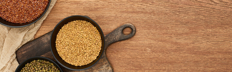 panoramic shot of bowls with grains and beans on wooden cutting board near canvas with copy space