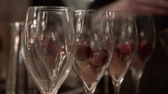 Bartender puts large grapes to empty champagne glasses at a fancy restaurant, close-up. Skillful barman making alcoholic cocktails in transparent glasses on bar counter under soft evening lighting.