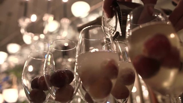 Bartender pouring cold champagne from iron shaker to tall glasses with large grapes at fancy restaurant, close-up. Skillful barman makes alcoholic cocktails on bar counter under soft evening lighting.