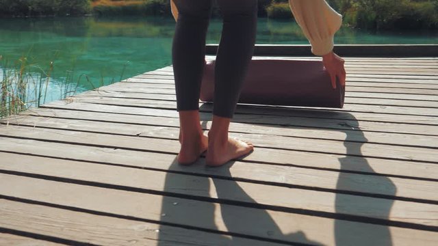 Close up shot od a young woman unfolding a yoga mat on a wooden path and starting with a mountain yoga pose. A beautiful lake surrounds her - calm and relaxing atmosphere.