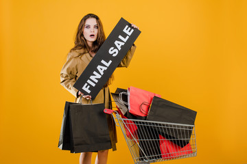 young woman in trench coat with final sale sign and colorful shopping bags in cart isolated over yellow