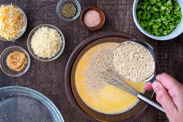 Ingredients for quinoa egg bake, raw egg mix in glass bowl with metal whisk, quinoa, cheese, herbs, broccoli, glass baking dish, on a wood table