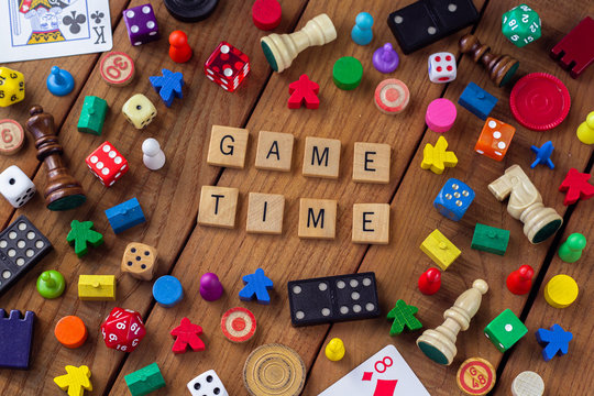 "Game Time" spelled out in wooden letter tiles, surrounded by dice, cards, dominoes, chess pieces and other game pieces on a wooden background