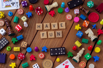 "Have Fun" spelled out in wooden letter tiles, surrounded by dice, cards, dominoes, chess pieces and other game pieces on a wooden background