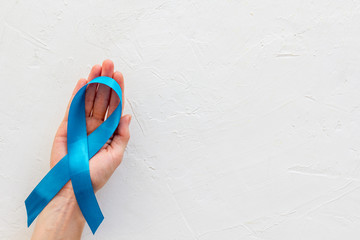 Blue ribbon in hands as symbol disease control on white background top view copy space