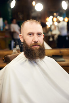 Close-up portrait of man brutal client with white cape sitting in barber chair in professional salon. Serious man with long brown beard looking in camera. Blurred background.