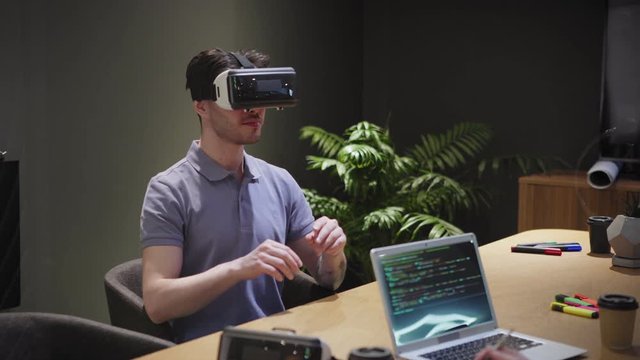 Camera panning left to man in VR headset interacting with virtual reality while sitting at table near female developers