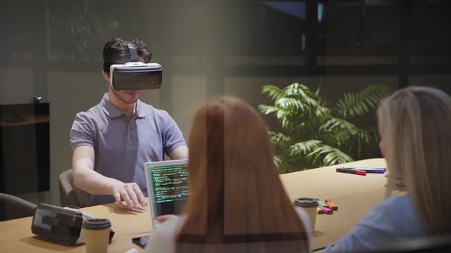 Camera tilting down to female programmers and male colleague conducting virtual reality app test while sitting at table with laptop in office