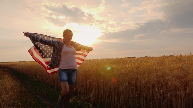 A woman with a USA flag runs across a wheat field in the sun's rays at sunset