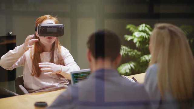 Camera tilting down to young female in VR goggles gesturing and exploring virtual reality while sitting at table near developers in office