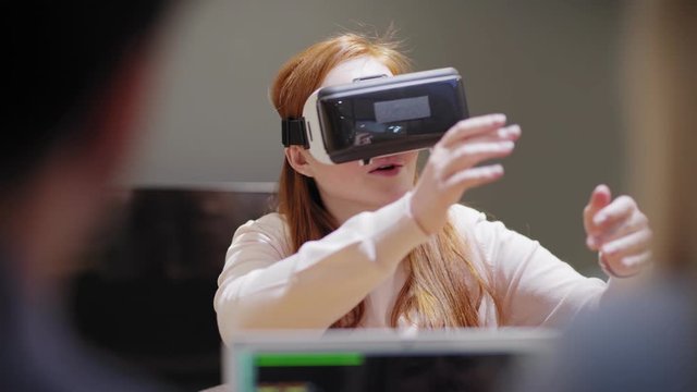 Young woman in VR headset interacting with virtual reality app while sitting near communicating programmers in office