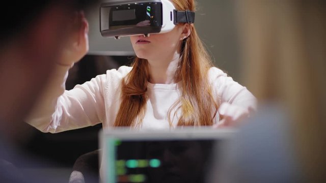 Camera tilting up from laptop with code to young woman exploring virtual reality near developers during VR app test in office