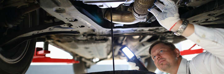 Male worker holding in hands electric torch inspecting oil leak under car portrait. Auto occupation concept