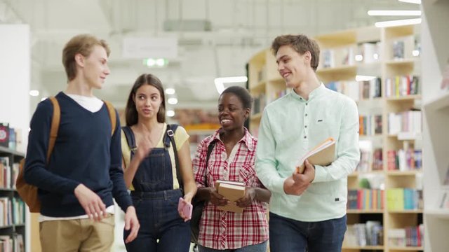 Group of four cheerful multiethnic students walking along book shelves in college library and having fun talking joyfully
