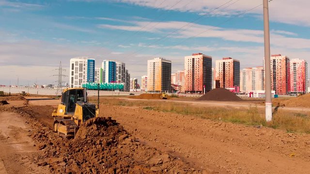 Flight back past the working bulldozer that flattens the ground for building a road in a new residential area. In background of a lot of building sand, earth and new residential area. Taken by drone
