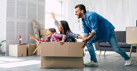 Little girl is riding in a cardboard box and pointing straight ahead while her mother imitates a...