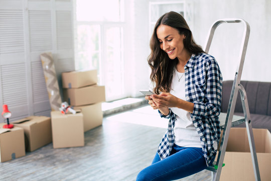 A small break. Young charming girl in a plaid shirt with long chestnut hair is sitting on the ladder surrounded by boxes, having a break after furnishing her new apartment and using her smartphone.