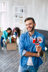 Some work to do. Handsome man is holding a scotch tape in his left hand while his wife and daughter are packing their belongings and preparing to move into the new house.