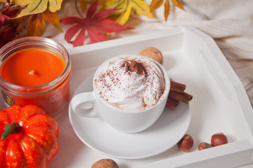 Cup of hot creamy cocoa with froth on the white tray with autumn leaves and pumpkins on the background