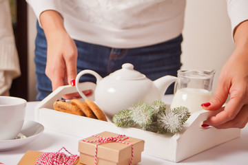 Woman holding a tray with teapot, cookies and jug of milk. Cup of tea, candy canes, Christmas gift boxes lies nearby on the white background