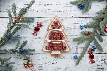 a lot of small red gift boxes in the form of a Christmas tree on a white wooden background, fir branches on the edge of the frame with Christmas toys. The concept of Christmas composition.