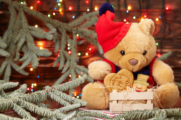 Christmas composition. Teddy bear in a red hat holding a box of homemade cookies on the background of defocused lights Christmas garland on the branches of the Christmas tree. brown wooden background