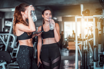 Fototapeta na wymiar Two young attractive Caucasian woman talking together and using smart phone or cellphone while drinking water and relaxation after hard workout at gym