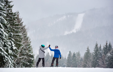 Back view of couple of snowboarders giving high five to each other, preparing descending from high...