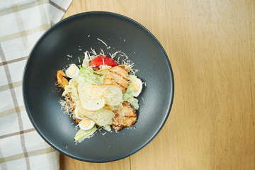 Caesar salad with croutons, parmesan, chicken, egg in a black stylish bowl. Restaurant serving. Selective focus, wooden background