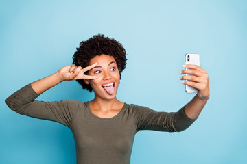 Photo of cute cheerful nice attractive black skinned woman fooling before camera showing v-sign sticking out wearing green sweater isolated over vibrant blue color background
