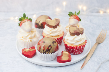 Cupcakes with cream cheese frosting decorated with fresh strawberries in white chocolate, chocolate red and golden balls and red chocolate hearts on the white plate. String lights on background.