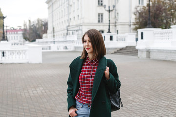 Fototapeta premium Young stylish brunette woman with short haircut wearing blue jeans, red plaid shirt, green coat, black backpack walking through the city streets. Trendy casual outfit. Street fashion