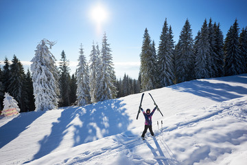 Happy smiling woman in helmet taking her ski in hands raised up and standing in middle of snow-covered mountain slope. Sunny day during winter vacation. Pine forest on background. Front general view.