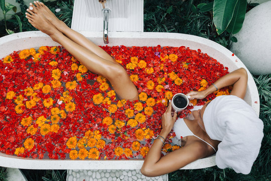 Woman Relaxing In Outdoor Bath With Flowers In Bali Spa Hotel.