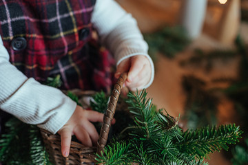Cute little girl is sitting by the Christmas tree on the floor in the room. Near the baby is a gingerbread house and a basket with fir branches.