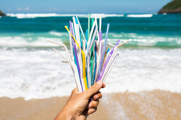 Fototapeta na wymiar Hand holding heap of used plastic straws on background of clean beach and ocean waves. Plastic ocean pollution, environmental crisis. Say no plastic. Single-use plastic waste