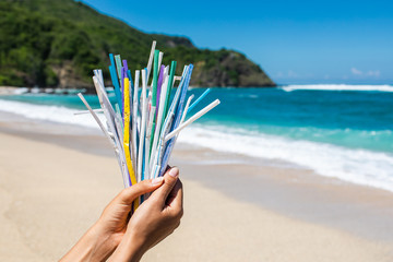 Hand holding heap of used plastic straws on background of clean beach and ocean waves. Plastic ocean pollution, environmental crisis. Say no plastic. Single-use plastic waste