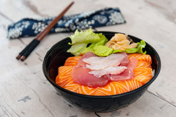 Japanese Chirashi made with assortment of three raw fishes, salmon, tuna and cod fish served on white vinegary rice with Wasabi sauce and ginger slices on top in a bowl.