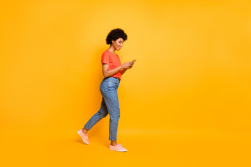 Side profile photo of cheerful cute attractive young influencer browsing through her telephone wearing jeans denim orange t-shirt sneakers isolated over vibrant color background