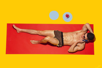 Top view of young caucasian male model's resting on beach resort on red mat and yellow background. Man's lying under sunlight and talking on phone. Concept of summertime, party, chill, vacation.