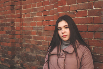 Naklejka premium Young cute pregnant girl in pink warm down jacket looks away. Late autumn, the cold is coming. Old brick wall on the background. toned photo.